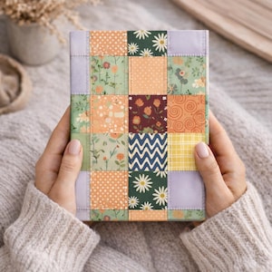 May include: A rectangular journal with a patchwork design. The cover features squares of various patterns, including floral, polka dots, and geometric shapes, in shades of green, orange, and blue. The journal is held by a person wearing a cream-colored sweater.