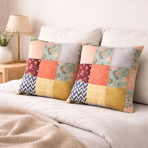 May include: Two square patchwork pillows with a variety of patterns and colors. The pillows feature squares of floral, geometric, and solid fabrics in shades of orange, green, yellow, and blue. The pillows are on a bed with a neutral-colored blanket.