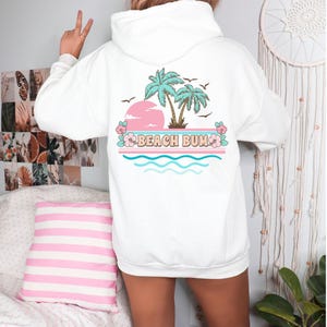 May include: White hoodie with a graphic on the back. The design includes a pink sun, palm trees, and the words "BEACH BUM". Blue and pink waves and flowers are also featured, with birds flying above. A casual, comfortable sweatshirt.