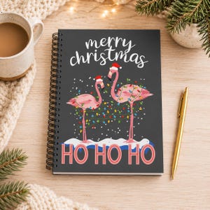 May include: A spiral-bound notebook with a dark gray cover featuring two pink flamingos wearing Santa hats, wrapped in colorful Christmas lights. The text reads merry christmas at the top and HO HO HO in large pink letters at the bottom.