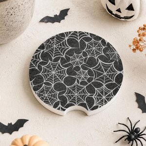 May include: A round car cup holder coaster featuring a black background with a repeating white spider web pattern. The coaster has a small semi-circular notch cut out from the bottom edge, designed for easy removal from a vehicle cup holder.