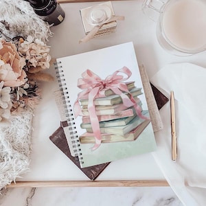 May include: A spiral-bound notebook featuring a watercolor illustration of a stack of books tied with a pink ribbon. The books have pastel-colored covers. The notebook rests on a stack of other books and a marble surface.