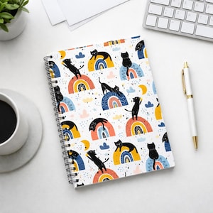 May include: Spiral-bound notebook with a whimsical pattern of black cats lounging on colorful rainbows, clouds, and moons. The notebook has a white background and a black spiral binding. A pen and keyboard are visible in the background.