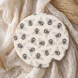 May include: A round, off-white car coaster featuring a repeating pattern of black and white bee illustrations. The coaster has a small notch for easy removal from a car cup holder. The design is simple and elegant.