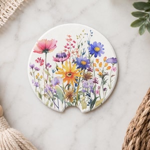 K&ouml;nnte beinhalten: Ein runder, wei&szlig;er Keramik-Auto-Untersetzer mit einer farbenfrohen Aquarell-Illustration verschiedener Wildblumen, darunter rosa, gelbe, blaue und violette Bl&uuml;ten. Der Untersetzer hat eine kleine halbkreisf&ouml;rmige Aussparung am unteren Rand.