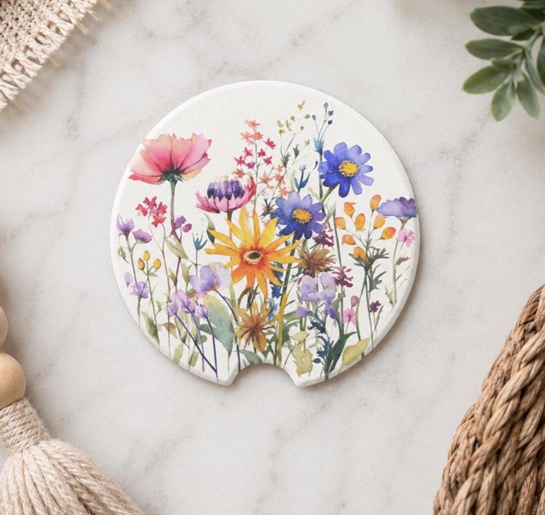 K&ouml;nnte beinhalten: Ein runder, wei&szlig;er Keramik-Auto-Untersetzer mit einer farbenfrohen Aquarell-Illustration verschiedener Wildblumen, darunter rosa, gelbe, blaue und violette Bl&uuml;ten. Der Untersetzer hat eine kleine halbkreisf&ouml;rmige Aussparung am unteren Rand.