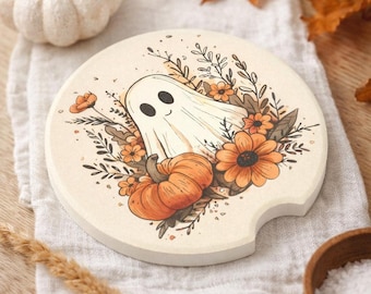 May include: A round car coaster with a cream-colored surface featuring a central illustration of a white ghost, an orange pumpkin, and various orange and brown flowers with dark brown line art details. The coaster has a small notch on the side.