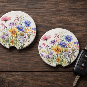 K&ouml;nnte beinhalten: Zwei runde, wei&szlig;e Keramik-Auto-Untersetzer mit einem farbenfrohen Aquarell-Druck von Wildblumen, darunter rosa, blaue und gelbe Bl&uuml;ten. Jeder Untersetzer hat eine kleine halbkreisf&ouml;rmige Einkerbung an der Unterkante. Ein schwarzer Autoschl&uuml;ssel liegt daneben.
