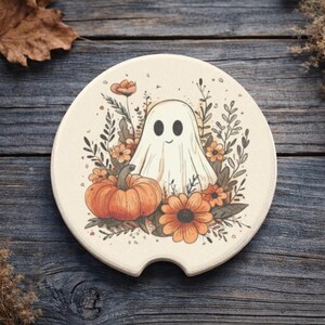May include: A cream-colored car coaster featuring a friendly ghost, pumpkin, and orange flowers. The design is surrounded by green foliage. The coaster has a small notch for easy removal from the car cup holder. Halloween themed.