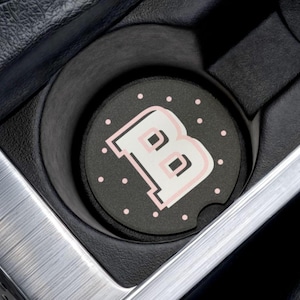 May include: A black car coaster with a white and pink letter "B" design. The coaster is surrounded by small pink polka dots. The coaster is in a car cup holder, with a silver trim.