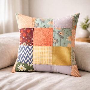May include: Square patchwork pillow with a variety of patterned fabric squares in shades of orange, red, blue, green, and yellow. The pillow has a soft, quilted appearance and is placed on a bed.