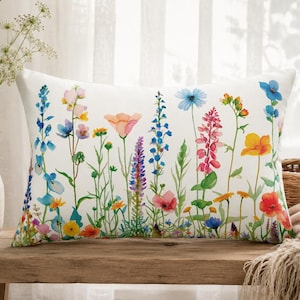 May include: A rectangular throw pillow featuring a white background with a colorful watercolor print of various wildflowers including blue, pink, yellow, and orange blossoms with green stems and leaves arranged in a meadow-like pattern.