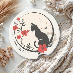 May include: A cream-colored car coaster featuring a black cat silhouette surrounded by red flowers and butterflies. The design is set within a black circular frame. The coaster is designed to fit in a car's cup holder.