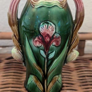 May include: A ceramic vase featuring a glossy dark green glaze with a raised, embossed iris flower design in shades of pink and red. The vase has two curved handles extending from the top, a scalloped rim, and green leaf accents.