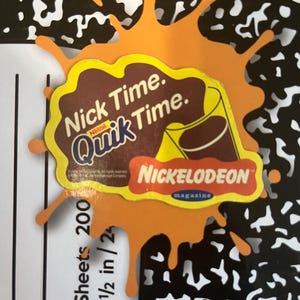 Puede incluir: Una forma naranja troquelada que se asemeja a una salpicadura, con una pegatina marrón y amarilla con el texto "Nick Time. Quik Time." y una imagen de dibujos animados de una bebida de chocolate. Una pegatina más pequeña, roja y amarilla, dice "Nickelodeon magazine."