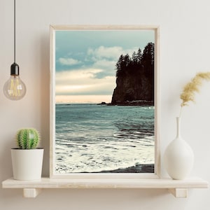 May include: A framed landscape print featuring a coastal scene. The image shows a rocky cliff with evergreen trees, the ocean, and a cloudy sky. The color palette includes shades of blue, gray, and brown, creating a serene and natural aesthetic.