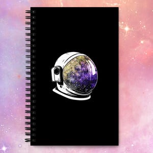May include: A black spiral notebook with a white astronaut helmet illustration. The helmet's visor is filled with a galaxy-like pattern of purple, yellow, and blue.