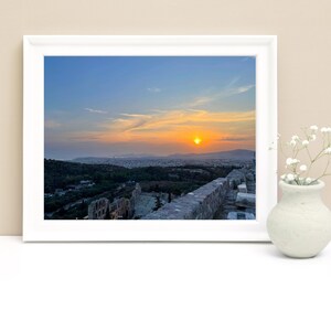 May include: A framed photograph captures a scenic sunset over a city, with a vibrant orange sun setting over the horizon. The foreground features a stone wall and a glimpse of ancient ruins. A small white vase with white flowers is on the right.