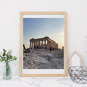 May include: A framed print of the Parthenon in Athens, Greece, at sunset. The image shows the ancient temple with its iconic columns, set against a clear blue and yellow sky. People are visible on the rocky terrain.