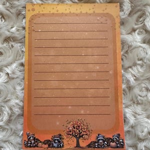 May include: A brown notepad with lined paper and a fall-themed design. The design features raccoons, a tree with orange leaves, pumpkins, and acorns. The background is a gradient of orange and brown.