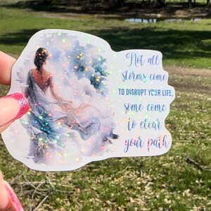 May include: A sticker with a watercolor-style image of a woman standing in a cloudy sky. The sticker has a glittery finish and features the text "Not all storms come to disrupt your life, some come to clear your path."