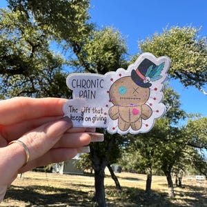 May include: A sticker with the text "CHRONIC PAIN" and "The gift that keeps on giving." It features a voodoo doll with a top hat, a heart, and stitched details. The sticker is held up against a backdrop of trees and a blue sky.