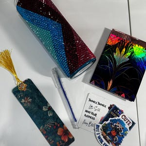 Puede incluir: Un conjunto de artículos coloridos que incluyen un vaso con un diseño geométrico, un marcapáginas con un diseño floral, una pequeña libreta con un diseño floral, un bolígrafo y una pegatina con un mapache y el texto "Soy lindo pero lucharé".