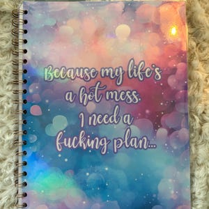 May include: Spiral-bound notebook with a colorful cover featuring a dreamy, bokeh-style background in shades of pink, blue, and purple. The text reads, "Because my life's a hot mess. I need a fucking plan..."