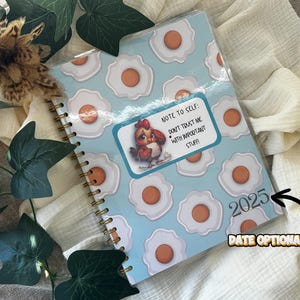 May include: A spiral-bound notebook with a light blue cover featuring a pattern of fried eggs. A sticker on the cover reads "NOTE TO SELF: DON'T TRUST ME WITH IMPORTANT STUFF!" and includes a cartoon chicken. The year 2025 is printed on the cover.