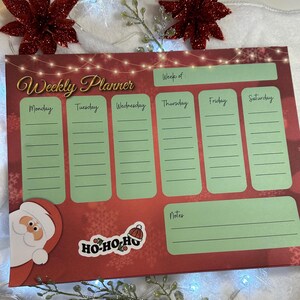 May include: A red and green weekly planner with a Santa Claus illustration and a "Ho-Ho-Ho" sticker. The planner has spaces for each day of the week, a notes section, and a space to write the week of.
