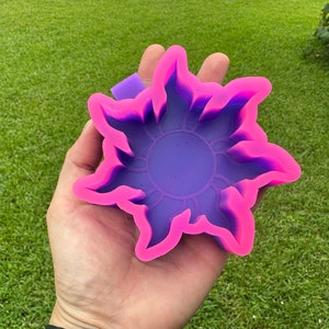 May include: A purple and pink silicone mold shaped like a sun with a raised design. The mold is held in a hand.
