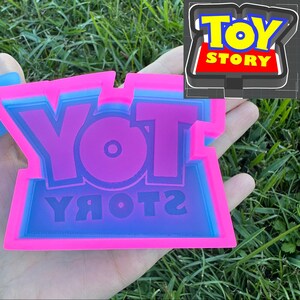 May include: A pink and blue silicone mold with the words "YOT YЯOTOЯ" in a reversed font. The mold is designed to create a replica of the Toy Story logo.