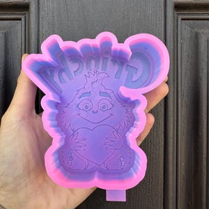 May include: A pink and purple silicone mold with the word "Grinch" and a cartoon image of the Grinch holding a heart.