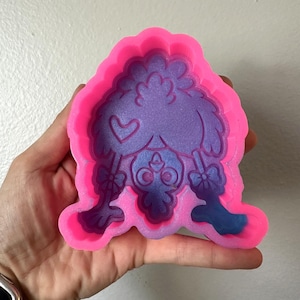 May include: A bright pink silicone mold featuring a recessed design of a stylized bird with a heart on its wing and bows on its legs. The interior of the mold is filled with a shimmering purple material, creating a contrast between the vibrant pink border and the glittery purple center.
