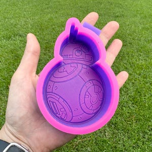 May include: A purple and pink silicone mold shaped like a Star Wars BB-8 droid. The mold is designed to create a 3D replica of the droid.