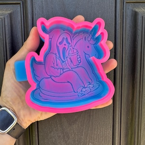 May include: A multi-layered silicone mold in shades of pink, blue, and purple. The mold features a cartoon image of a character from the Scream movie franchise riding a unicorn and drinking from a cup. The mold has a blue tab.