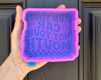 May include: A purple and blue silicone mold with the text "Run Car Not Mouth" in a wavy font. The mold has a checkered border and two checkered flags. The mold is held in a hand.