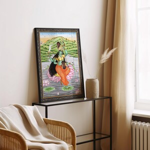 K&ouml;nnte beinhalten: Gerahmtes Kunstwerk, das eine t&uuml;rkisfarbene Gottheit zeigt, die auf einer rosa Lotusblume sitzt. Die Figur tr&auml;gt orangefarbene und gelbe Kleidung und h&auml;lt ein Schwert und einen Sch&auml;del. Der Hintergrund zeigt eine Landschaft mit einem Fluss und gr&uuml;nen H&uuml;geln. Der Rahmen ist schwarz mit einem dekorativen Rand.