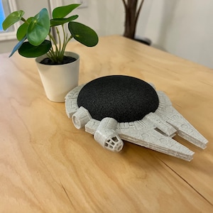 May include: A gray 3D-printed Millennium Falcon-shaped stand holds a black, round speaker. The stand is on a light wood surface, with a small potted plant in the background. The speaker is a smart home device.