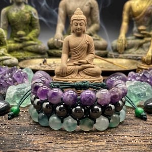Könnte beinhalten: Ein dreireihiges Perlenarmband bestehend aus einer Reihe violetter Amethystperlen, einer Reihe polierter schwarzer Hämatitperlen und einer Reihe blassgrüner Fluoritperlen, befestigt mit einer dunkelgrünen, verstellbaren Kordel.