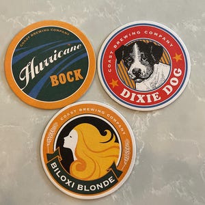 May include: Three round coasters from Coast Brewing Company. One coaster features the text "Hurricane BOCK" on a green and orange background. Another coaster has a dog illustration with the text "Dixie Dog". The third coaster has a woman's silhouette with the text "Biloxi Blonde".