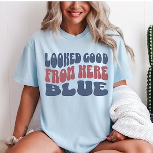 May include: A light blue t-shirt with the text "LOOKED GOOD FROM HERE BLUE" in a retro font. The text is in red and blue.