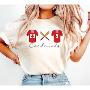 May include: Cream-colored t-shirt featuring a baseball-themed design. The design includes two red baseball jerseys with the name "KING" and numbers 23 and 9, a crossed baseball bat, and the word "Cardinals" in a script font.