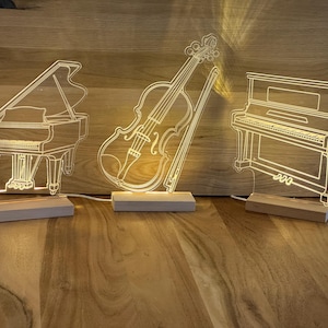 Könnte beinhalten: Drei beleuchtete Musikinstrument-Skulpturen: ein Flügel, eine Geige und ein Klavier. Jedes besteht aus klarem Acryl mit weißer Kontur, montiert auf einem Holzsockel. Die Instrumente sind von innen beleuchtet und erzeugen ein warmes Leuchten.