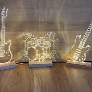 May include: Three illuminated musical instrument-shaped LED lights. The set includes a guitar, a drum kit, and a bass guitar, each mounted on a wooden base. The lights are white and cast a warm glow, perfect for a music-themed room.