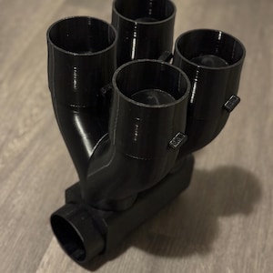 Könnte beinhalten: Ein schwarzer, 3D-gedruckter Verteiler mit vier zylindrischen Öffnungen oben und einer einzelnen Öffnung unten. Der Verteiler hat ein gebogenes Design, wobei die Öffnungen zur Mitte hin abgewinkelt sind. Das Objekt besteht aus einem glatten, matten Material.
