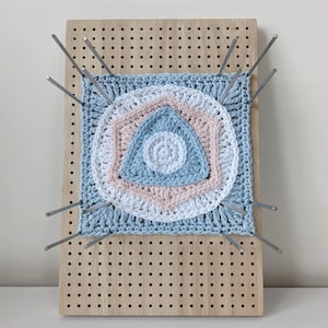 May include: A wooden pegboard with a crocheted mandala design in shades of blue and pink. The mandala is attached to the pegboard with metal pins.