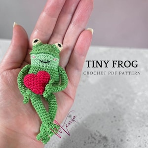 Puede incluir: Una rana verde de ganchillo hecha a mano sosteniendo un corazón rojo. La rana tiene ojos negros y una boca sonriente. El texto "TINY FROG CROCHET PDF PATTERN" se muestra en la imagen.