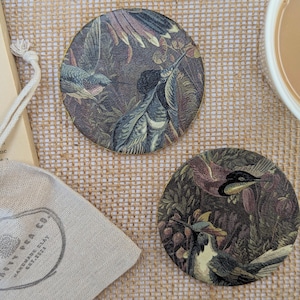 May include: Two circular coasters featuring a bird and floral pattern, with a gold edge. A small drawstring bag is visible, with the text "Crafty Pea Co. Handmade Clay Est. 2022". The coasters are made of clay.