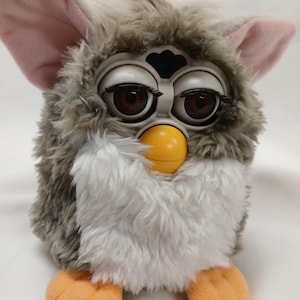 May include: A vintage Furby toy with grey and white fur, large brown eyes, and a yellow beak. The toy has pink ears and orange feet. A classic electronic toy from the 1990s.